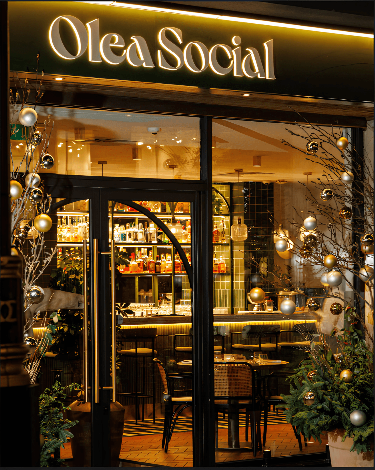 Olea Social shopfront at night, lit sign and windows decorated with gold and silver baubles